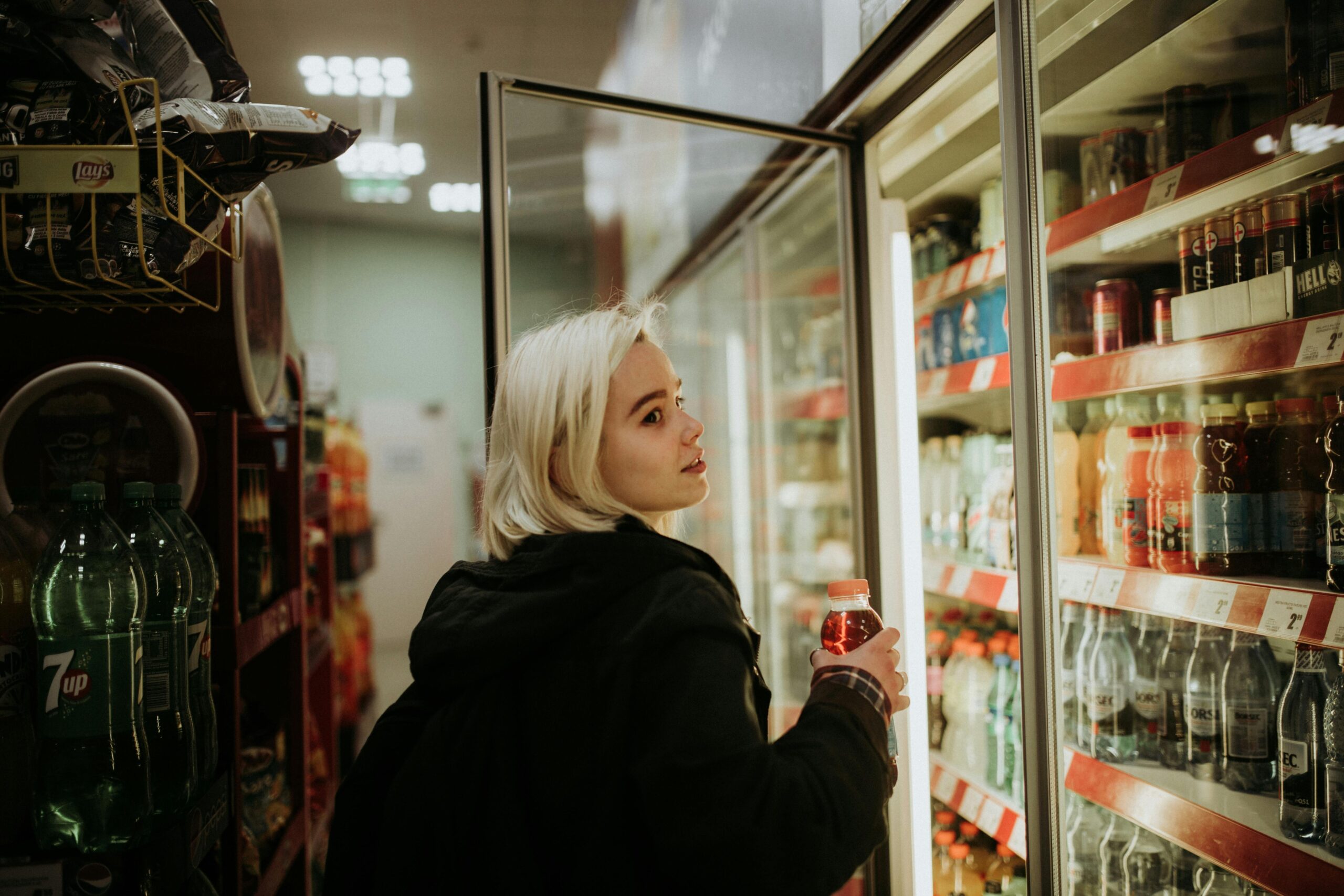 Blonde woman selecting a beverage from a refrigerated shelf at a convenience store.